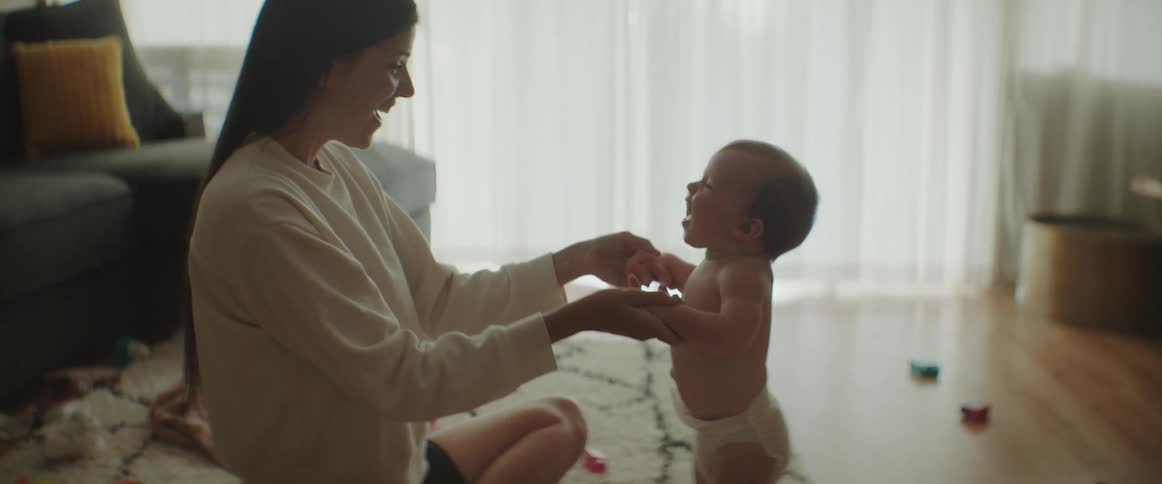 A woman is sitting on the floor helping her baby stand up