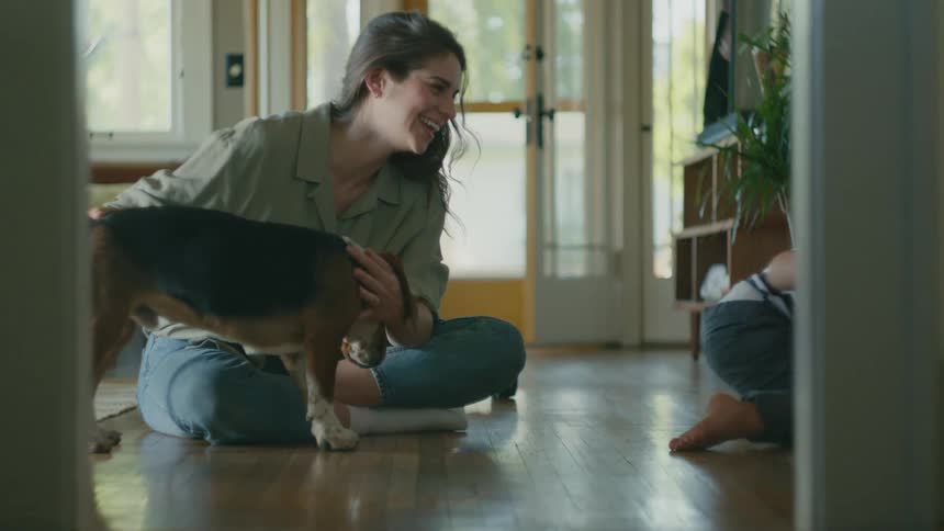 A woman is sitting on the floor petting a dog and a young boy is sitting next to her and he pets the dog and laughs as he scoots away