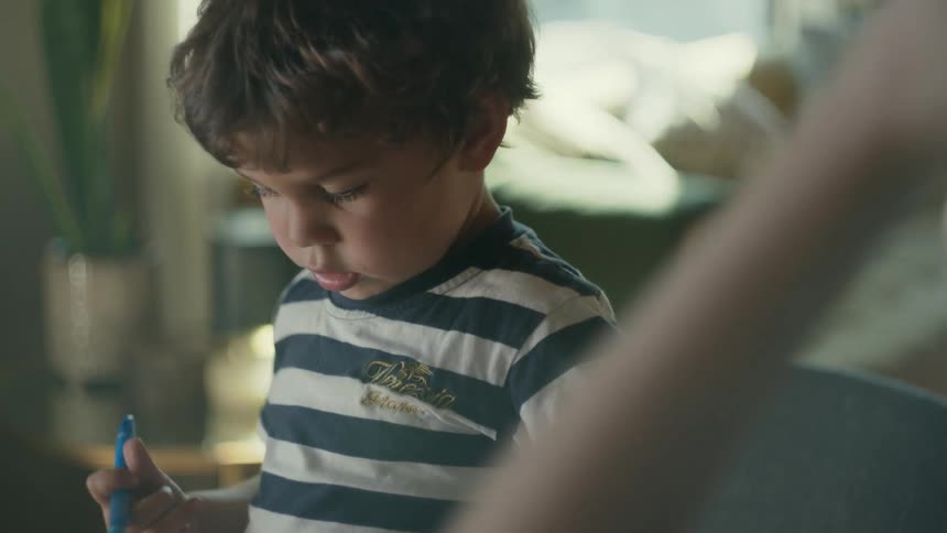 A boy collects markers before uncapping one and drawing on a sheet of paper while sitting at a table with a woman