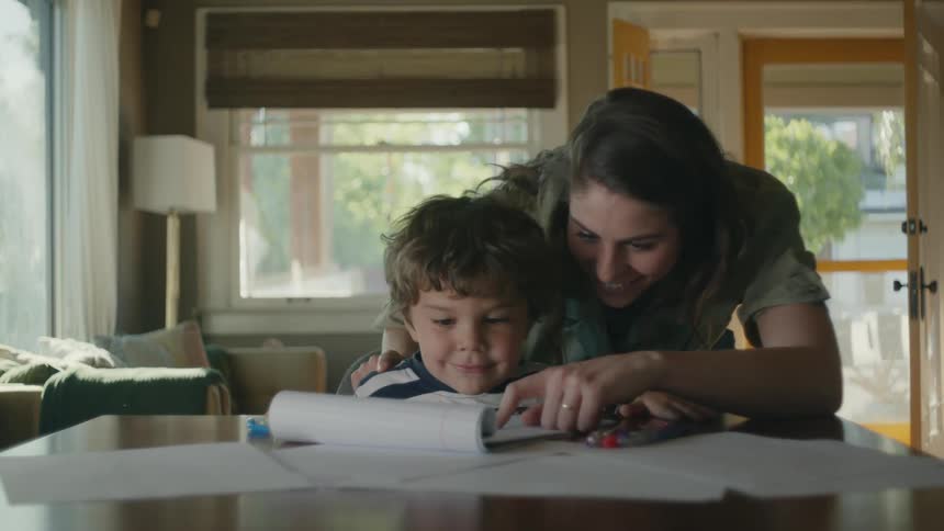 A little boy is sitting at a table drawing and his mother walks over to see his picture