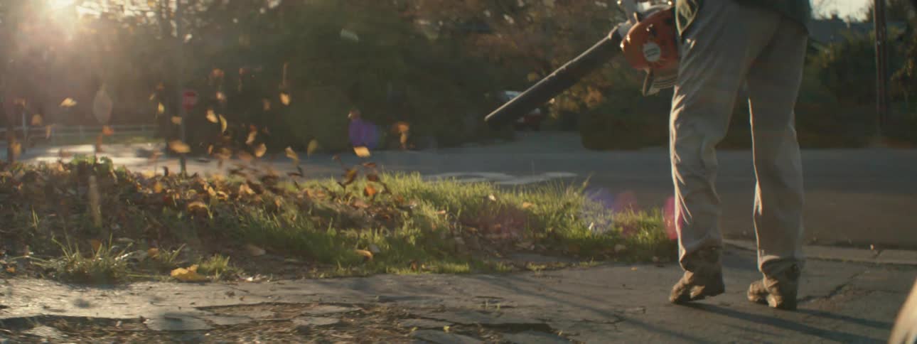 The sun shines brightly as a man is outside using a leaf blower outside in his yard