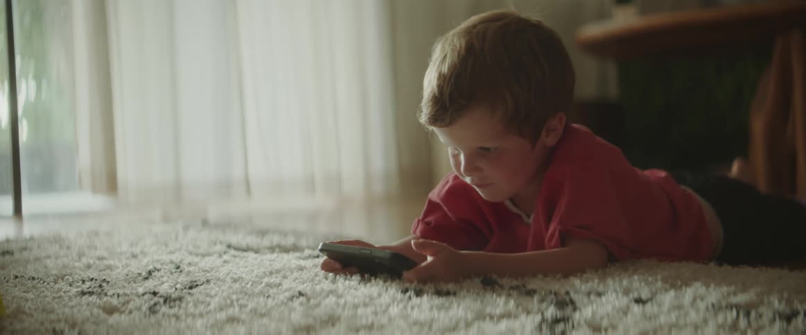 A young boy watches a video on a phone while lying on the carpet