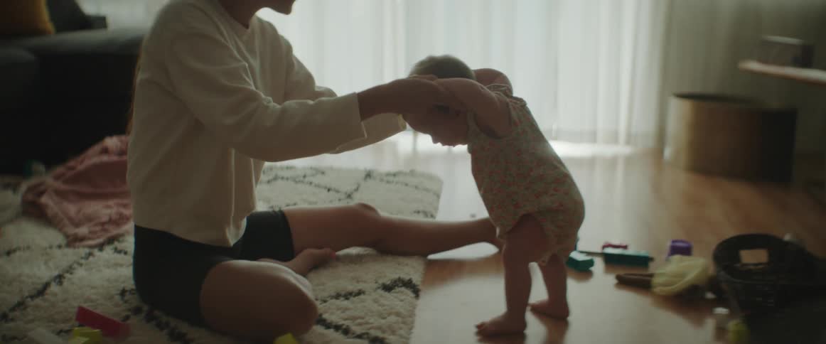 A woman helps a baby to stand while sitting on the floor of a living room during the day