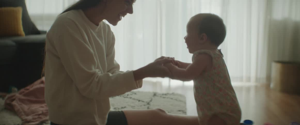 A woman sitting on the floor of a living room holds the hands of a baby as it attempts to stand