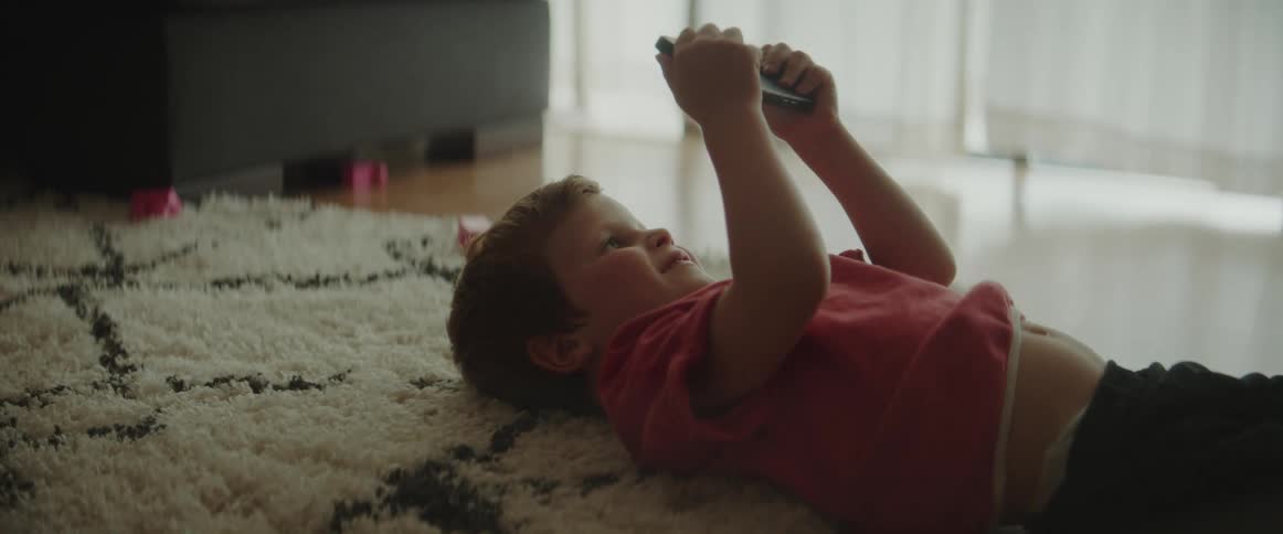 A little boy smiles while lying on the rug watching a video on a smart phone