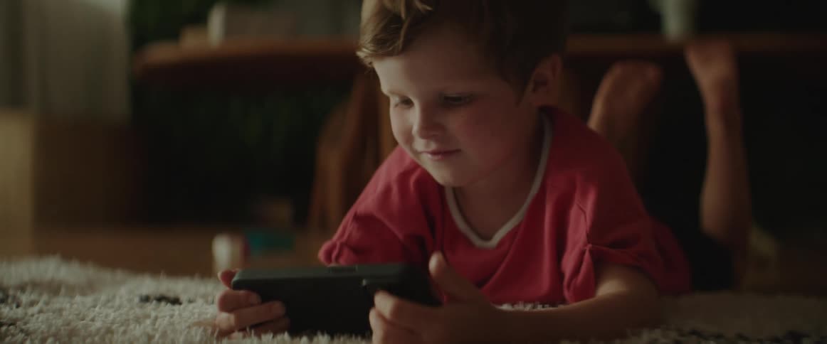 A little boy is lying on a rug on the floor holding a cell phone in his hands and smiling as he looks at it