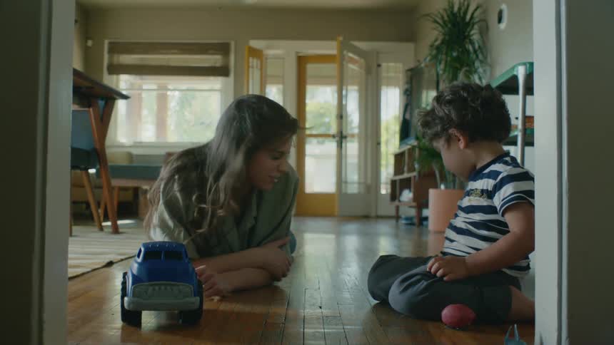 A woman is lying on a hardwood floor talking to a little boy playing with a toy car