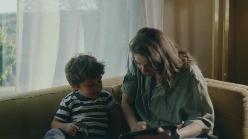 A little boy is sitting on a couch playing with a slinky and a woman sitting next to him is showing him a tablet