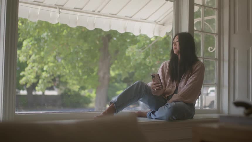 A woman is sitting on a window ledge holding her phone as the wind blows the leaves of a tree outside the window