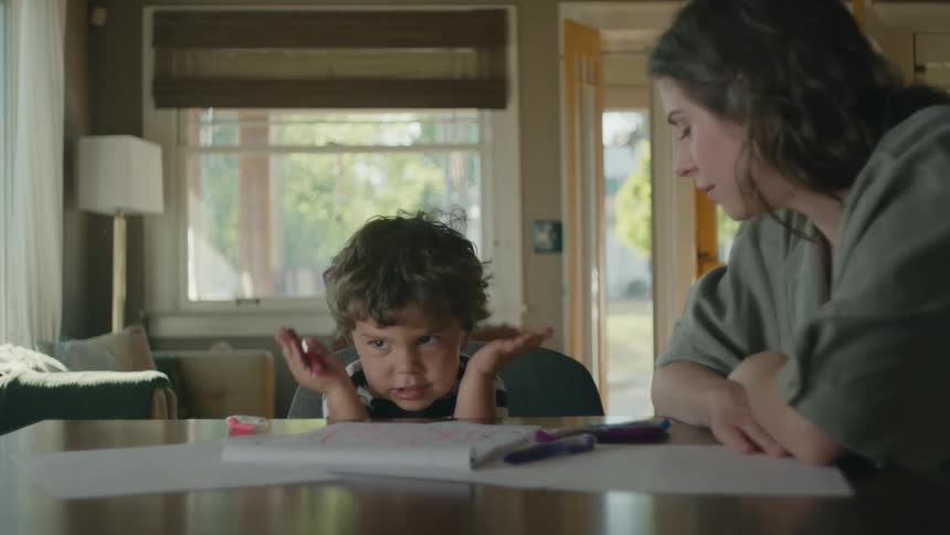 A little boy uses his arms hands and body while telling a story to his mom while he colors
