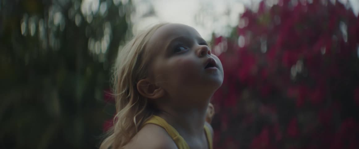 A little girl with blond hair is playing outside and there are flowers and trees behind her