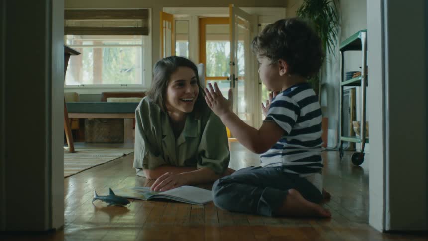 A woman is lying on a hardwood floor with her hand on an open book talking to a little boy sitting on the floor near her