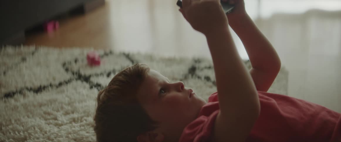 A young boy is lying on a rug on the floor holding up a cell phone as he looks at it