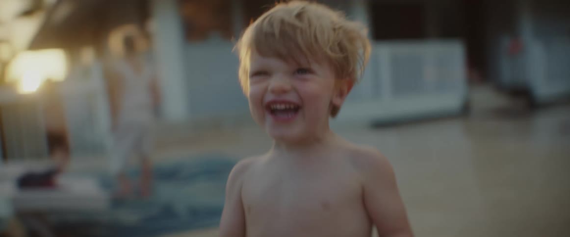 A shirtless child chases the camera around a balcony in the evening
