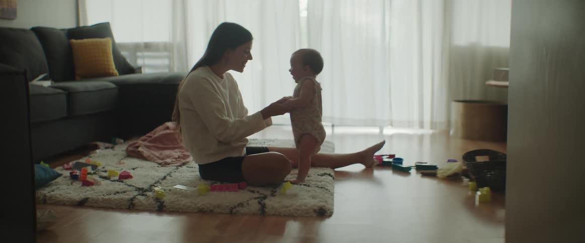 A womans and holds her baby up by its arms while they sit on the floor