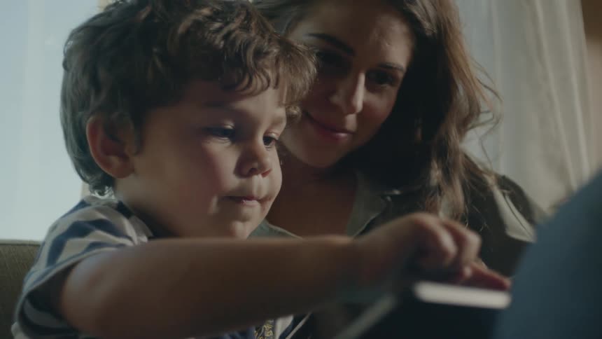 A young boy sits with his mom at home and plays on a tablet as she watches
