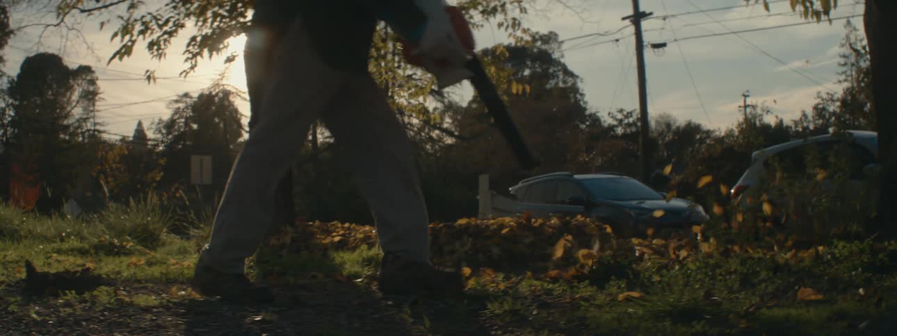 A man is outside blowing leaves across his yard with a leaf blower as the sun shines brightly in the distance
