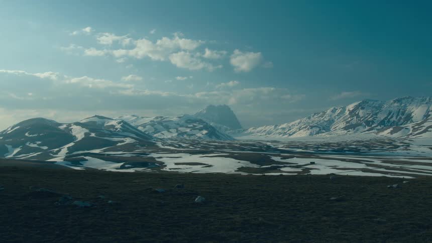 Melted boulders made of snow dot a field below a mountain range