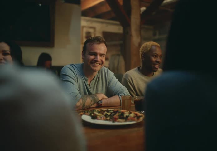 A group of men and women sit at a table with pizza and drinks in restaurant and talk and laugh