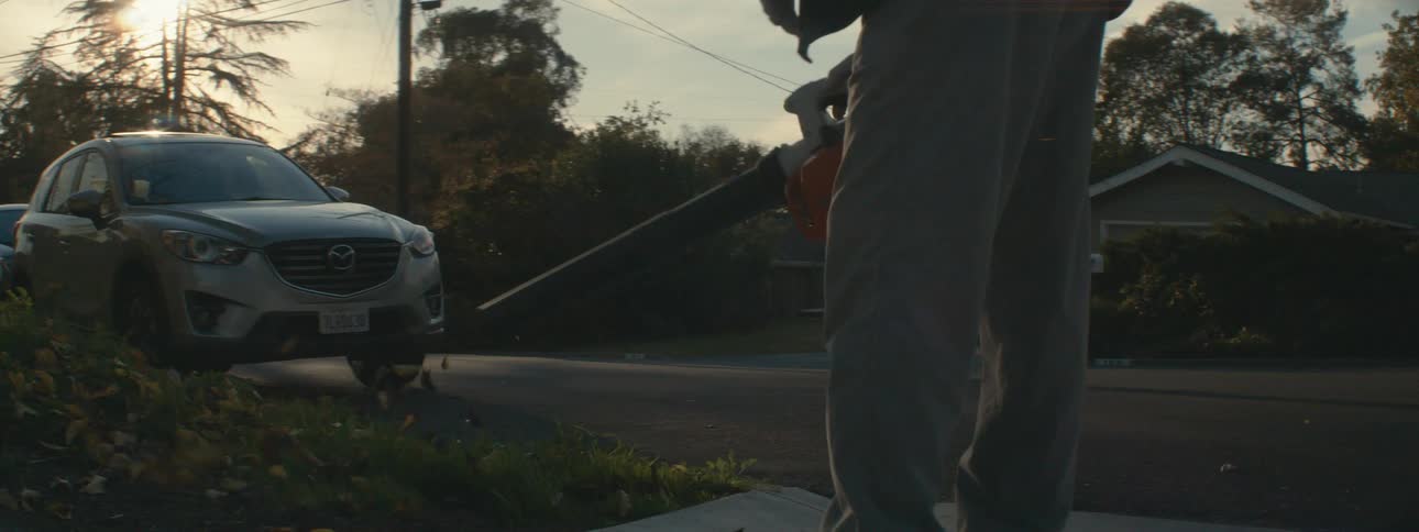 A man is standing outside in his yard holding a leaf blower as he starts it and blows leaves across his yard