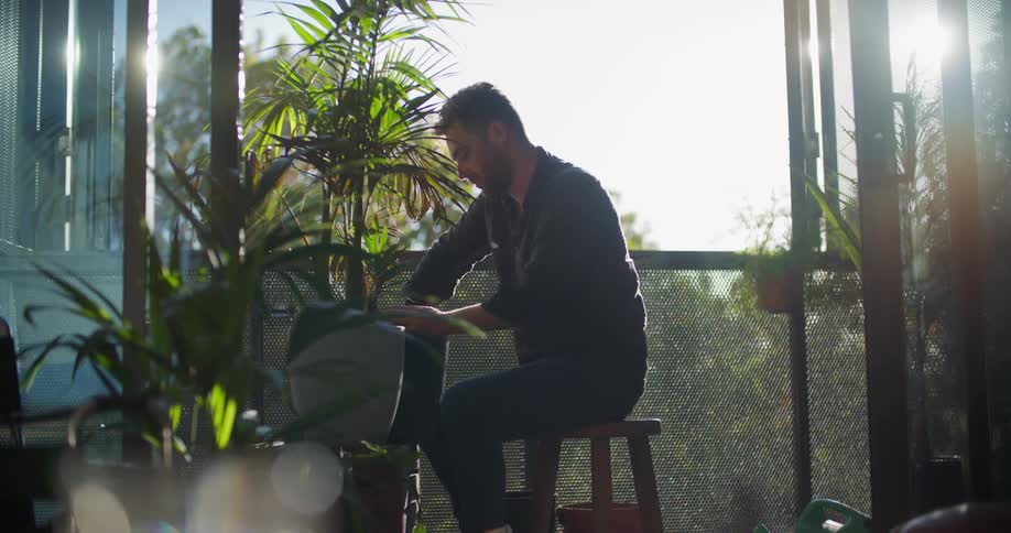 A man is sitting outside putting soil around a plant in a pot with a garden trowel