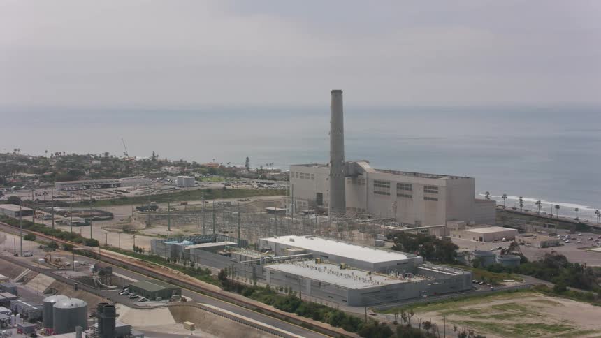 Tall smoke stack at a factory building and cranes along the shore as waves roll in under a cloudy sky