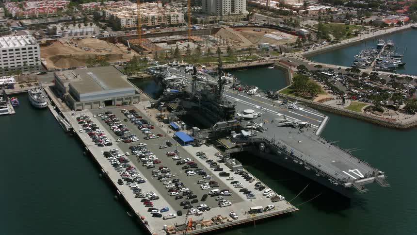 Airplanes are on an aircraft carrier docked along the coast and many cars are in a parking lot on shore