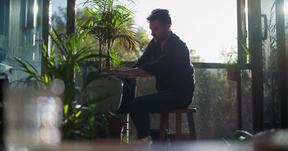 A man is sitting outside on a stool putting soil around a plant in a pot