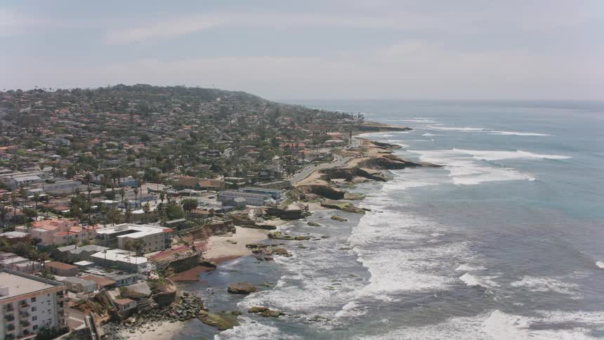 An aerial view of the ocean with waves rolling in to the shore that is lined with hotels and cars driving in the distance past a neighborhood