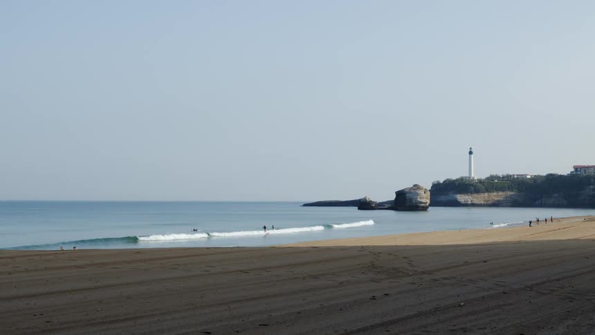 Waves roll over the beach as people are in the water and on shore and there is a lighthouse in the distance