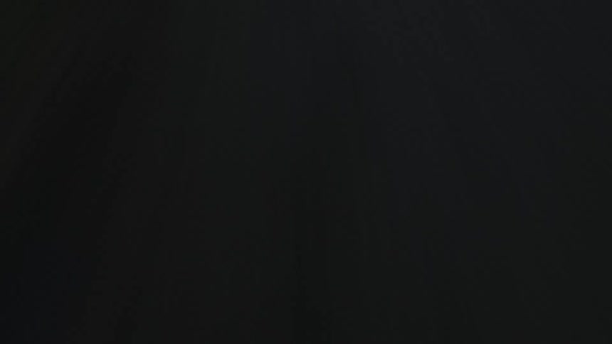 A white automobile is traveling on a dark forest road with the headlights on