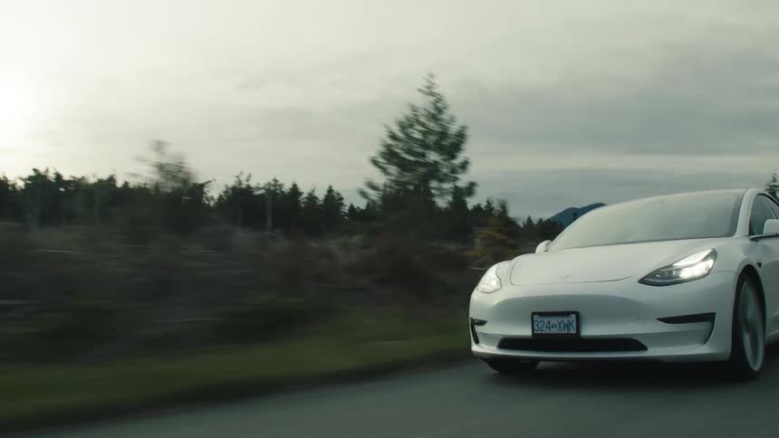 A white automobile is traveling on road surrounded by forests under a cloudy sky