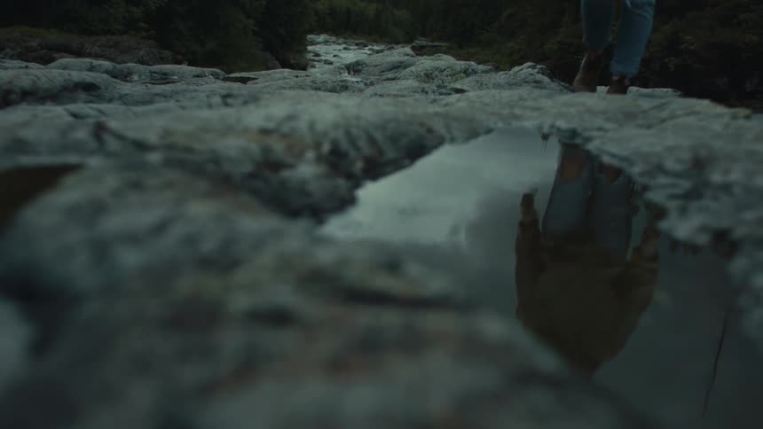 Reflection on a puddle of a woman walking over the rocks on a pine tree forest
