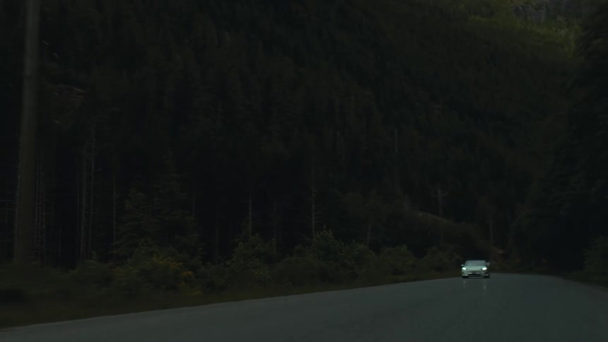 A white automobile is traveling on a forest road with the headlights on