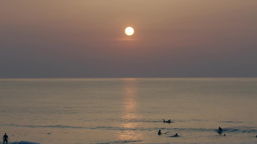 A group of people swim and surf in the ocean under a low sun