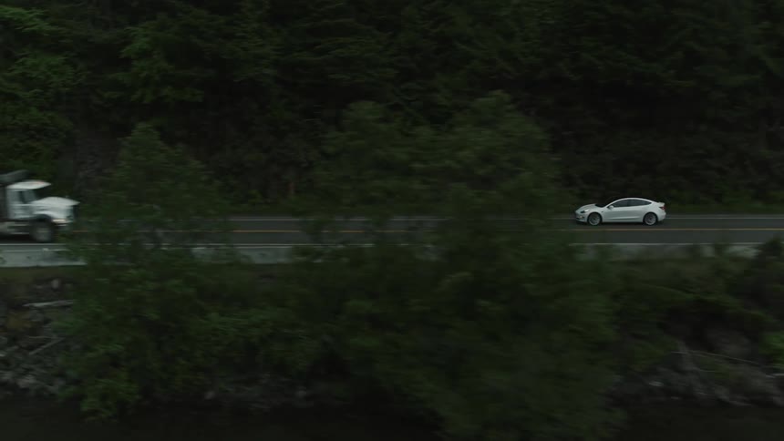 A car is driving on a rural highway near a wooded area