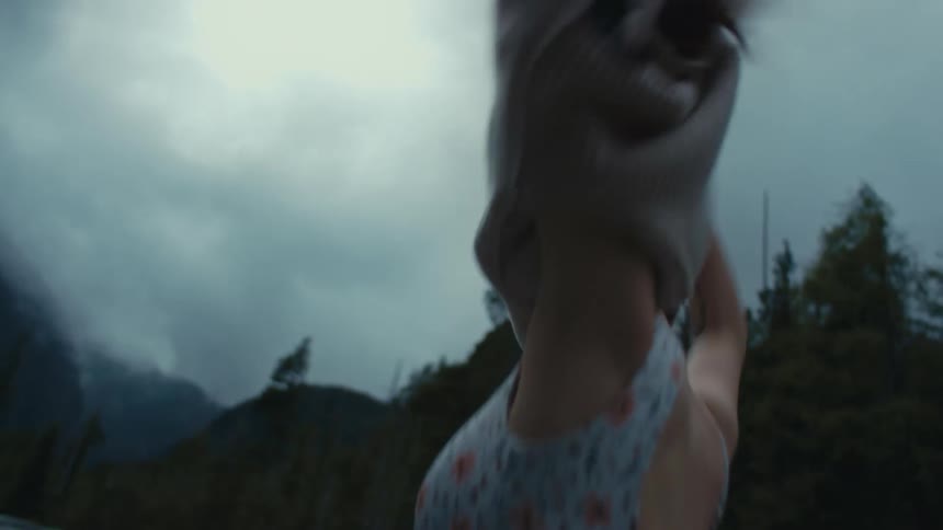 Woman strips off sweater revealing a bikini while standing on bank of lake with forest and mountains in the background