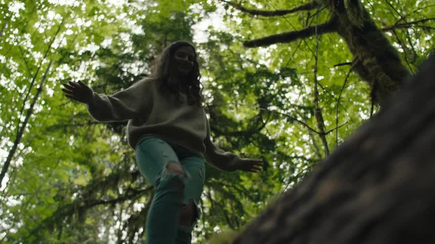 Woman balances carefully while walking along large moss covered fallen tree in forest