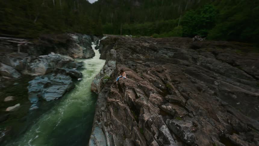 A teenage girl is climbing on rocks above river rapids in a forest under a cloudy sky