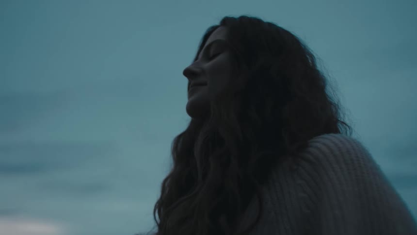 Woman wearing sweater stands on shore and closes her eyes to breath deeply