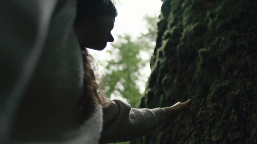 A teenage girl puts her hand on the moss covered bark of a tree trunk