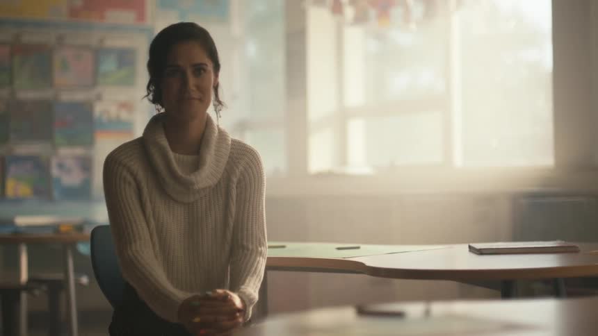 A woman is standing in a classroom and she sits down in a chair and smiles