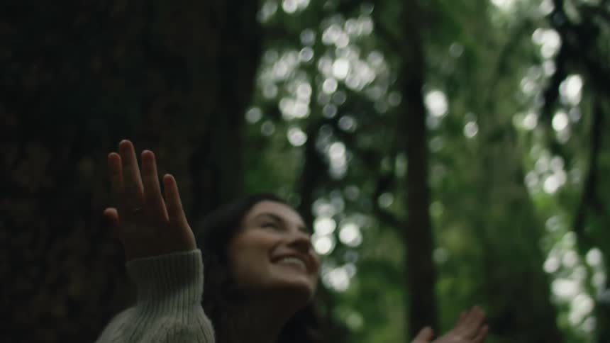 A woman holds up her hands and laughs as rain falls while she stands in the woods