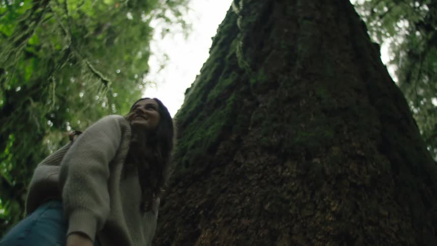 Woman wearing sweater stands at base of large moss covered tree then looks around