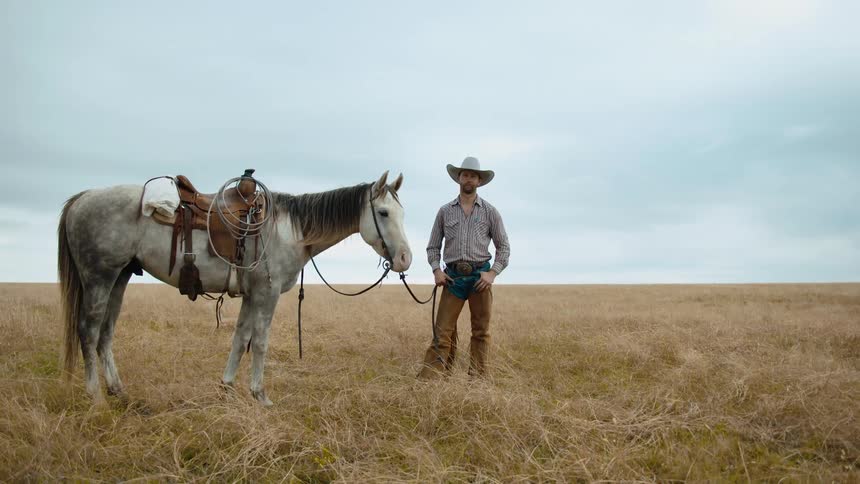 Cowboy standing next to a horse on a pasture grassland looking at the camera