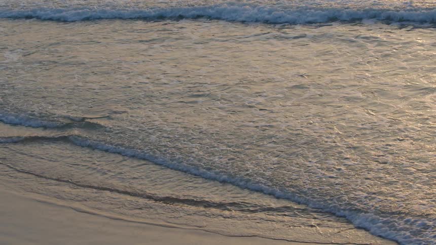 Sunlight reflects on the water as waves roll into shore and over the sand on a beach