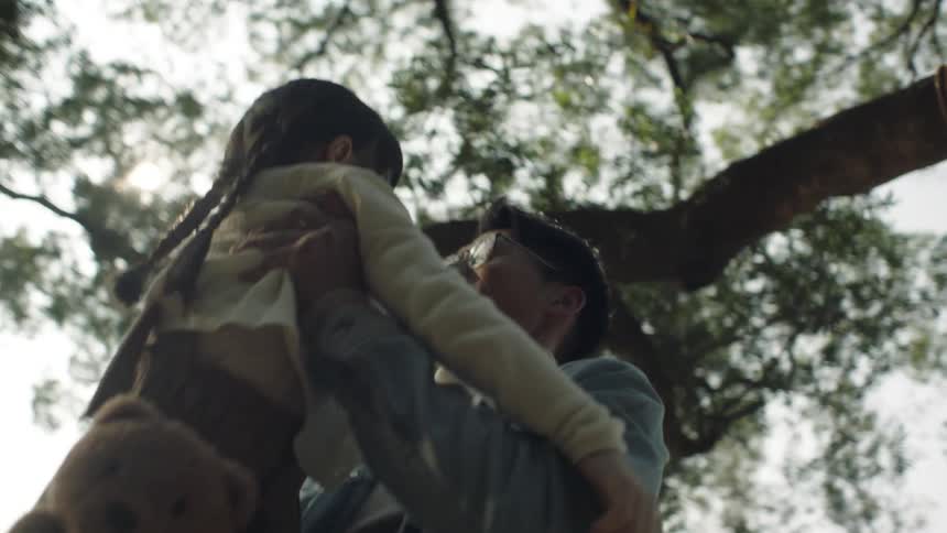 A man playfully lifts a young girl while standing outdoors under a tree during the day