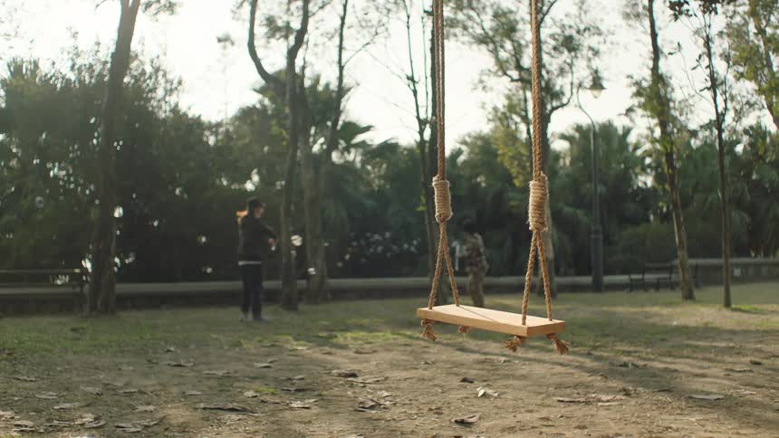 The sun shines on a swing in a park as children play with soap bubbles in the distance and a man walks by with a child and a dog