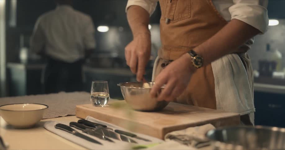 Chef stands in restaurant kitchen stirring mixture in metal bowl set on wooden chopping block