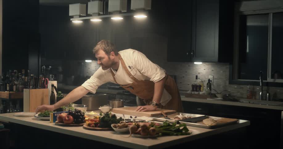 A male chef is reaching for a plate as he prepares food in the kitchen